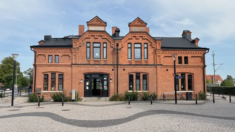 En röd tegelbyggnad med två våningar och flera fönster. Byggnaden har två ingångar och en symmetrisk fasad med dekorativa detaljer. Framför byggnaden finns en stenlagd yta med några bänkar och lyktstolpar.
